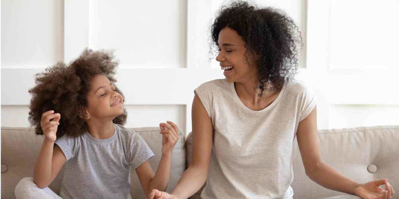 Hero image of a woman and a young girl laughing on a couch, sitting cross-legged with their hands in meditating position