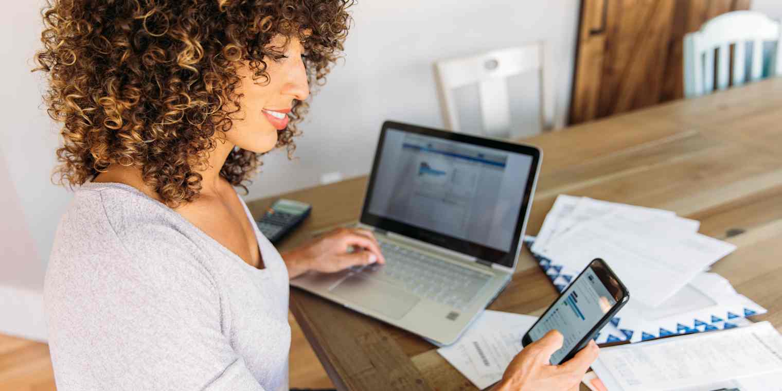 A woman uses a smartphone and computer while reviewing paperwork.