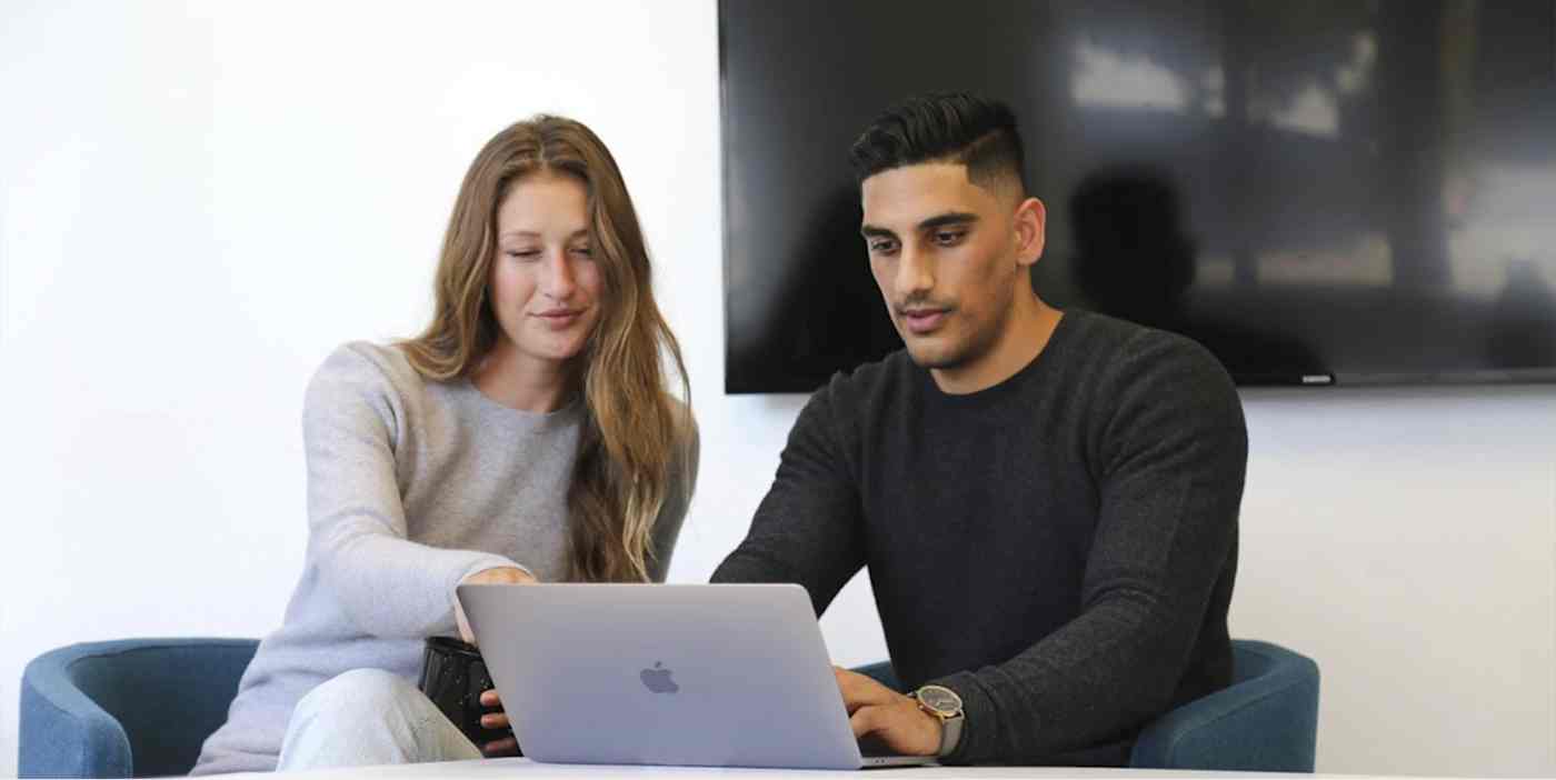 Hero image of two people sitting in modern chairs with a screen behind them, pointing to a computer