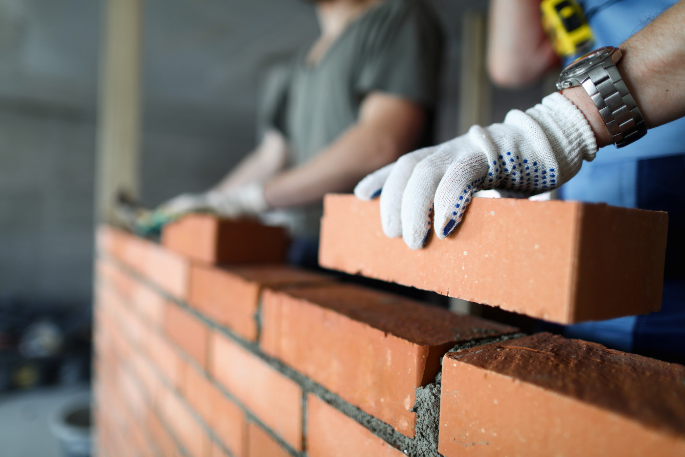 A brick wall being laid. The image shows a worker's hand in the foreground holding a brick above the in-progress wall.