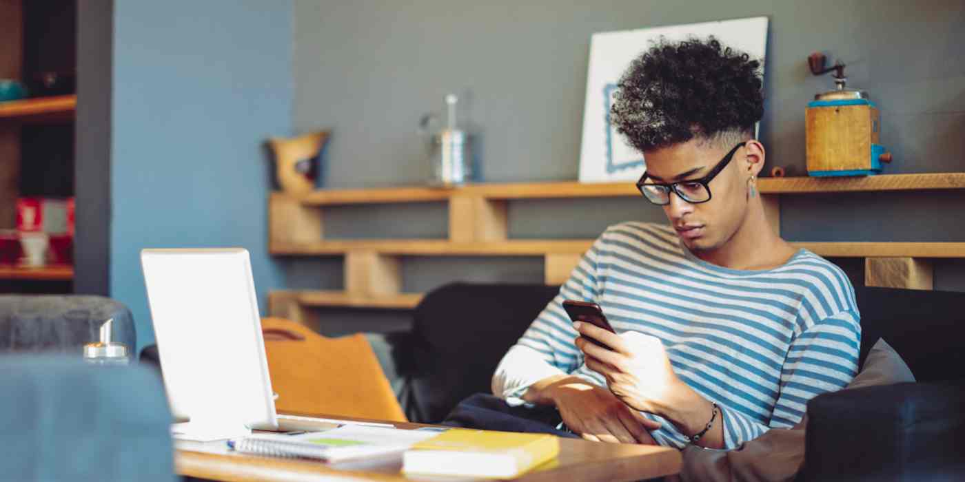 Hero image of a young man with glasses looking at his phone while in front of a computer