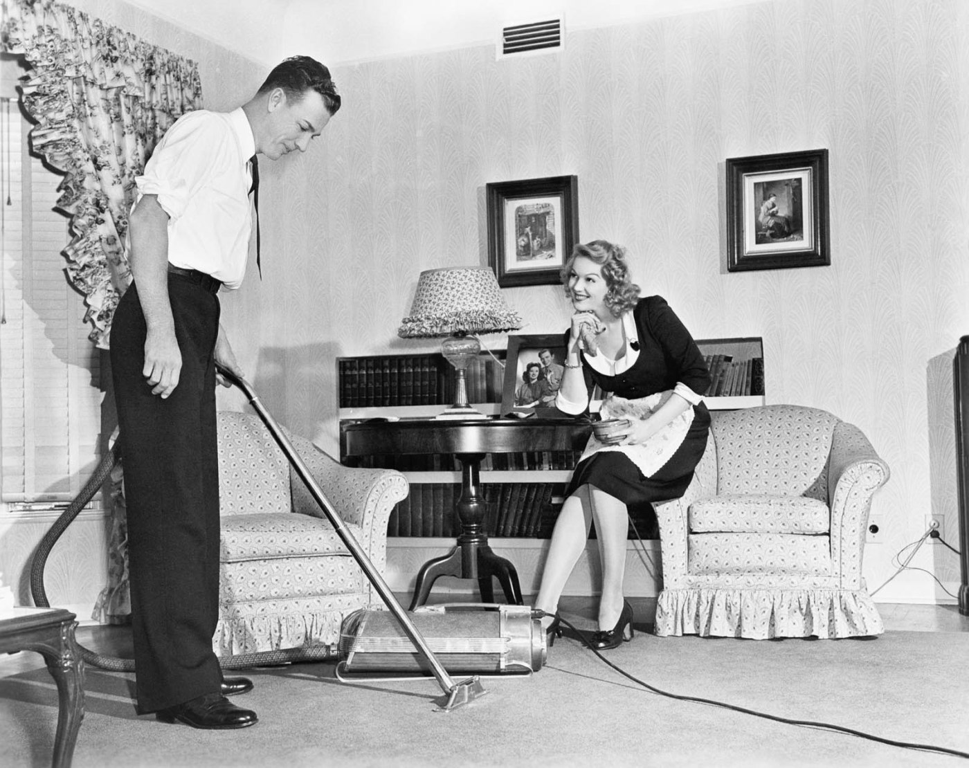 Photo of a 1950s style advertisement with a man vacuuming a living room floor while a woman watches from a chair.