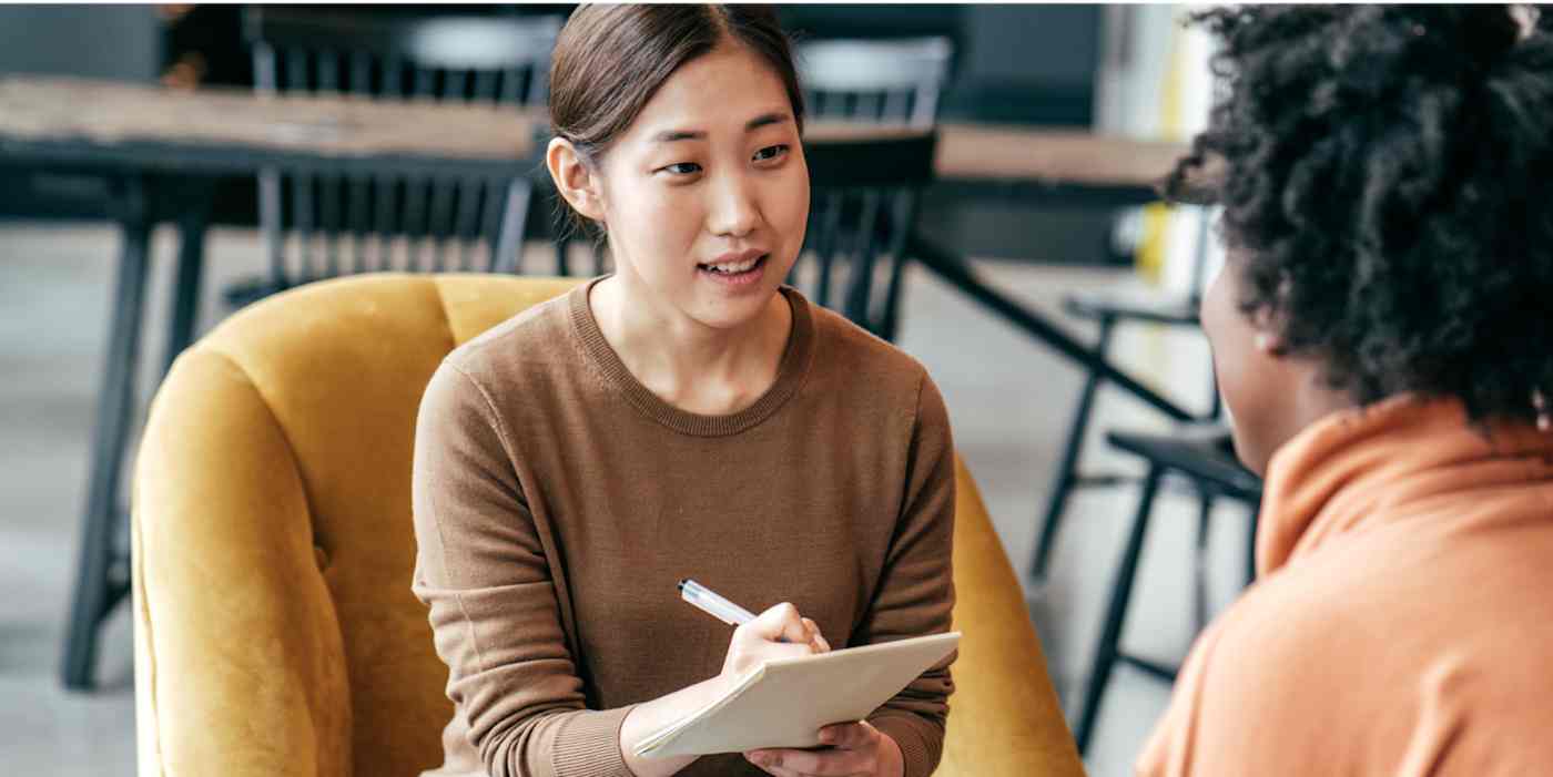 Screenshot of a woman interviewing another woman, holding a pad of paper and a pen