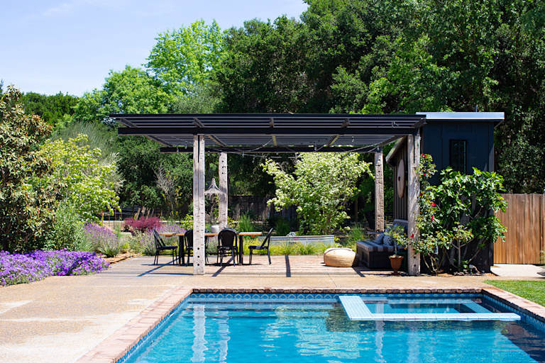A sunny day in a lush yard. There is a bright blue pool shining in the foreground, a covered patio with a black metal arbor behind it and lush green oak trees in the background. 