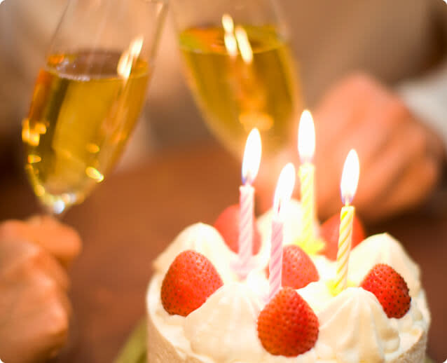 A birthday cake with candles; two hands holding glasses of sparkling wine in the background