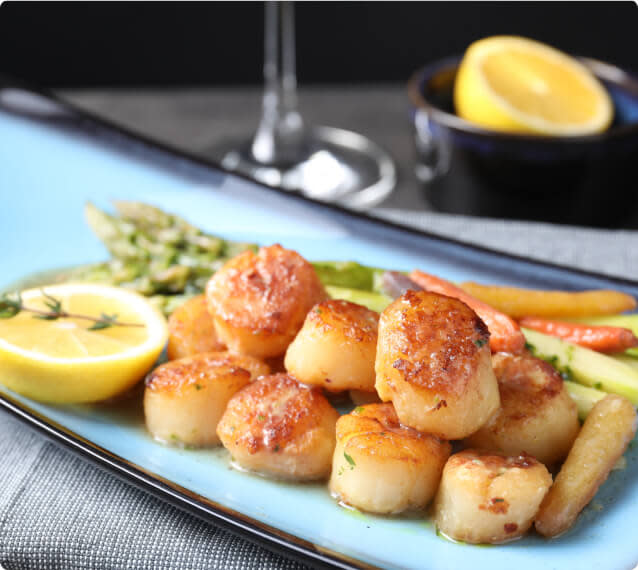 A plate of seared scallops, a lemon slice, and asparagus on a table with a wine glass in the background