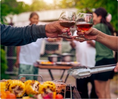Two people toasting with glasses of red wine near a grill at an outdoor barbecue