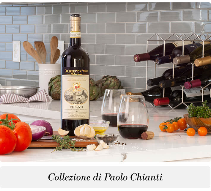 A bottle and two glasses of Collezione di Paolo Chianti red wine on a kitchen countertop with assorted vegetables and kitchen items in the background