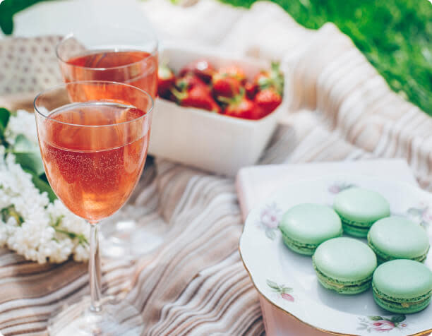 Two glasses of sparkling rosé wine on a picnic blanket next to a plate of pistachio macarons and a basket of fresh strawberries