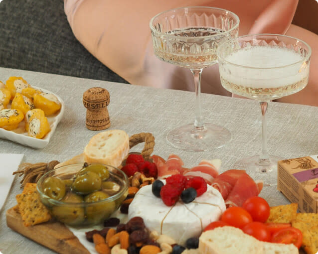Two glasses of champagne wine next to an assortment of cheeses, vegetables, charcuterie, nuts and olives