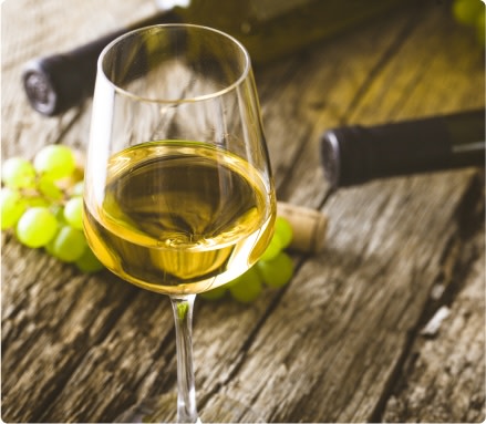 A glass of dry white wine on a table, next to some grapes, a wine cork and bottles