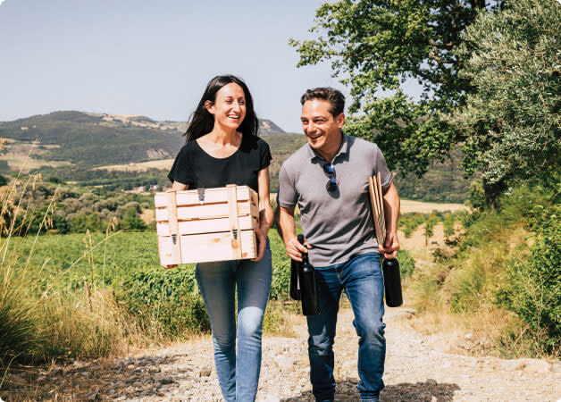 A woman and a man walking through a Tuscan vineyard holding a crate and bottles of wine