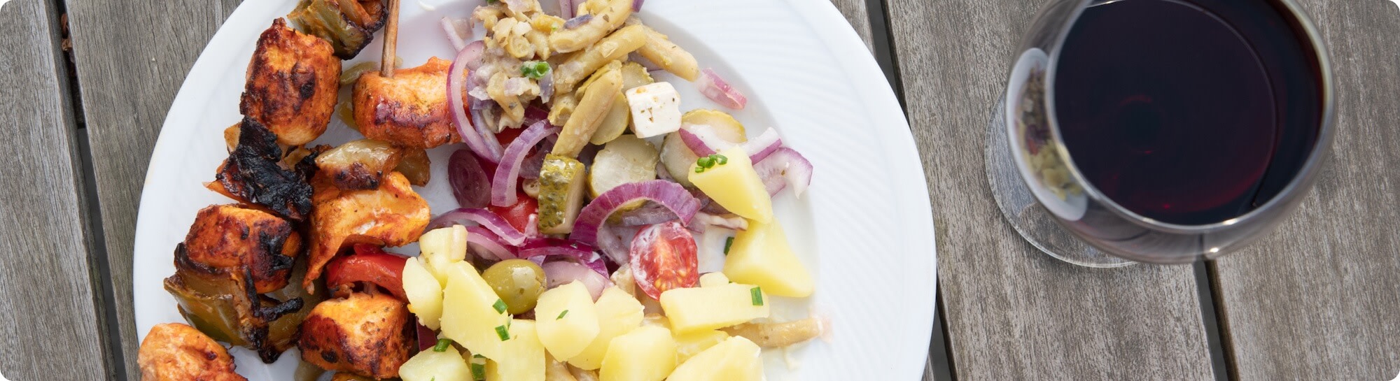 a glass of red Malbec wine next to a plate of food; grilled meat skewers, potatoes and salad