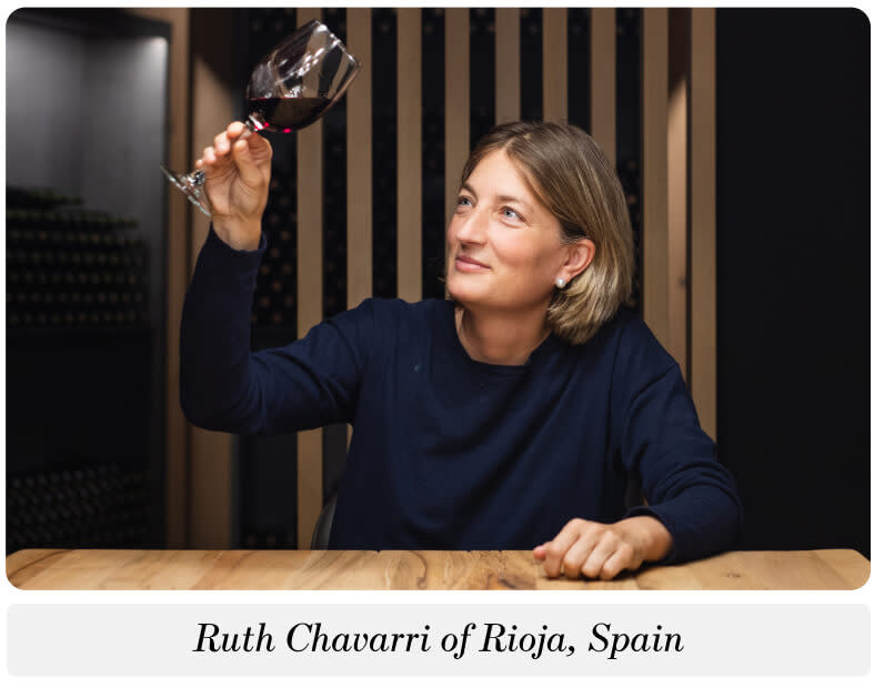 A photo of a woman sitting at a table holding up a glass of red wine to look at the color. The caption below reads: "Ruth Chavarri of Rioja, Spain"