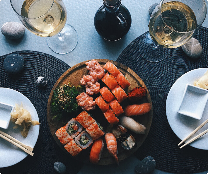 An overhead view of a dinner table set with a large plate of sushi, with glasses of white wine