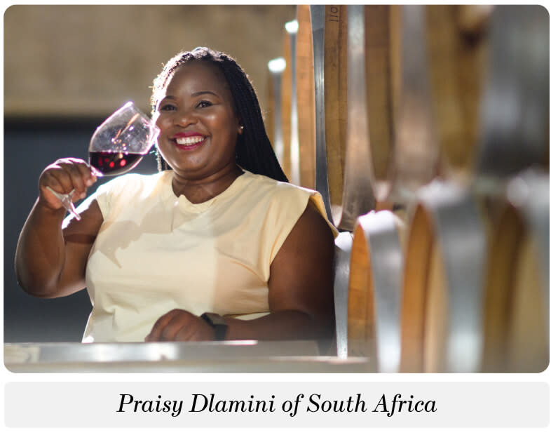 A woman standing next to barrels of wine holding up a glass of red wine and smiling. The caption below reads: "Praisy Dlamini of South Africa"