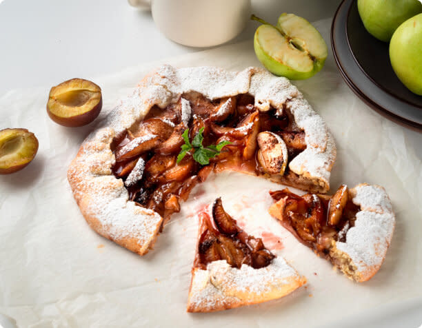 A plum galette with slices taken out, next to some cut plums and apples