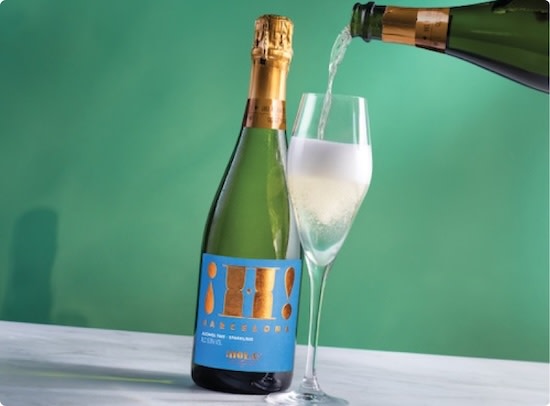 A bottle of ¡Hola! sparkling alcohol-free wine, with another bottle being poured into a champagne flute