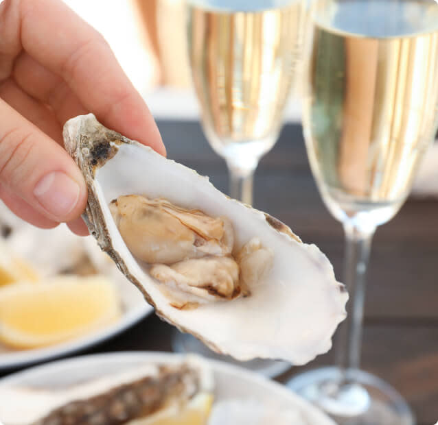 A hand holding a raw oyster on the half shell with two flutes of Champagne in the background