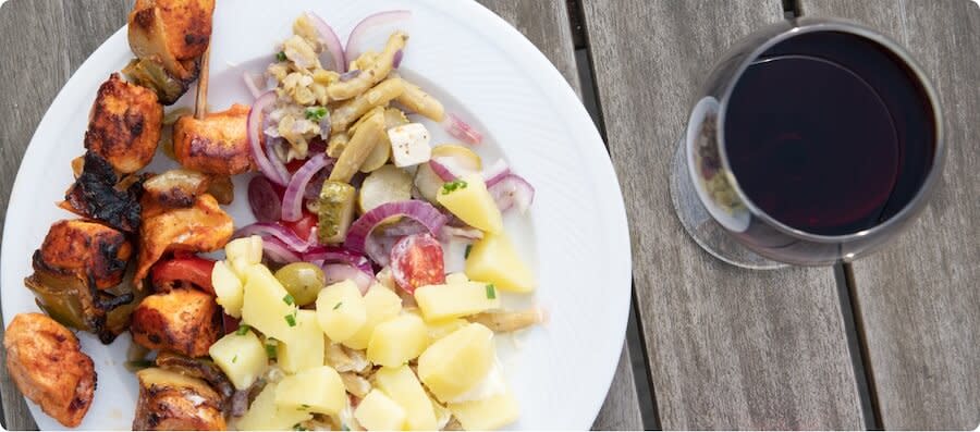 a glass of red Malbec wine next to a plate of food; grilled meat skewers, potatoes and salad