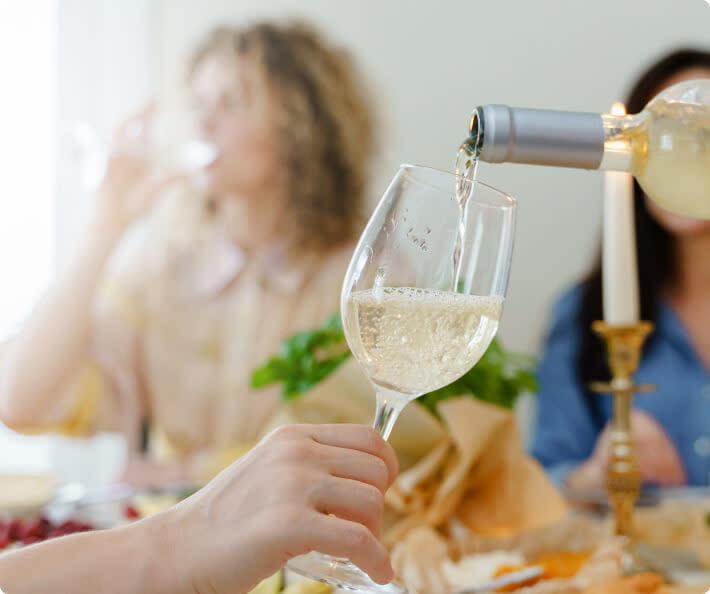 A close-up shot of someone pouring a glass of white Pinot Grigio wine, with blurred people in the background at a dinner table
