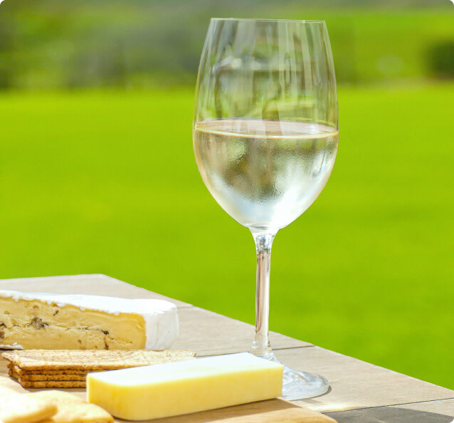 A glass of Pinot Grigio white wine on a table outdoors; there is a cheese board on the table next to the glass with slices of cheese and crackers