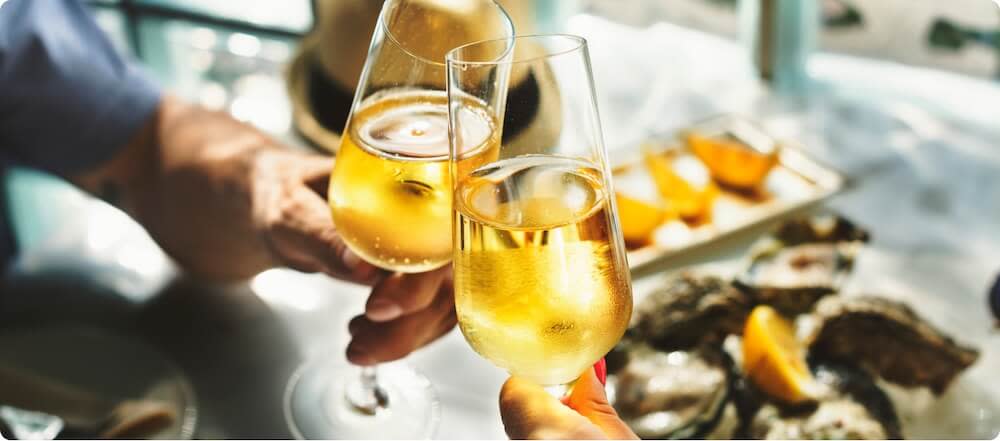 Two glasses of Champagne wine toasting above a table with foods such as oysters