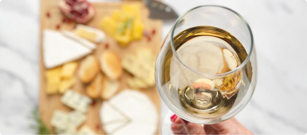 A hand holding a glass of Chardonnay wine with a cheese board in the background