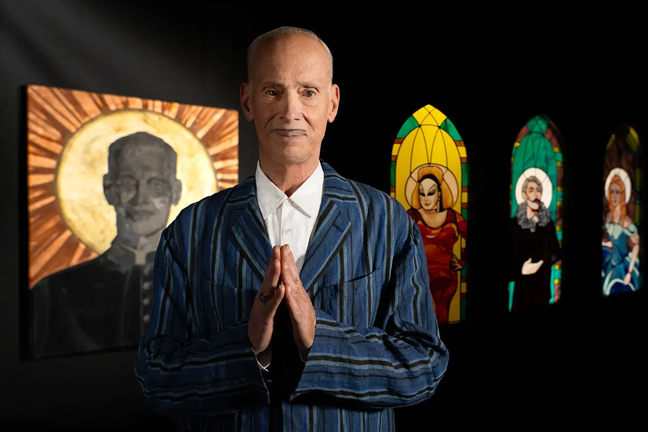 Image of John Waters in front of stained-glass windows in gallery