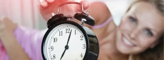 Bild på en väckarklocka med en kvinna i bakgrunden som sträcker sig för att stänga av väckarklockan.