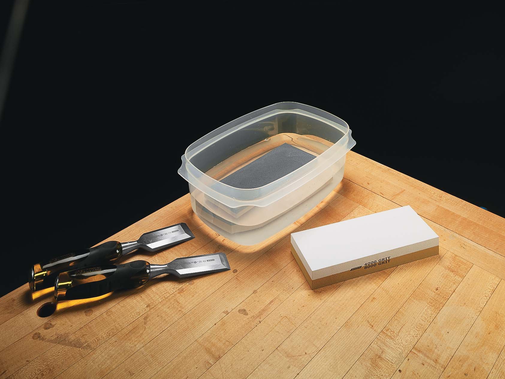 Image of tupperware container half filled with water, a man-made waterstone, and two silver wood carving chisels