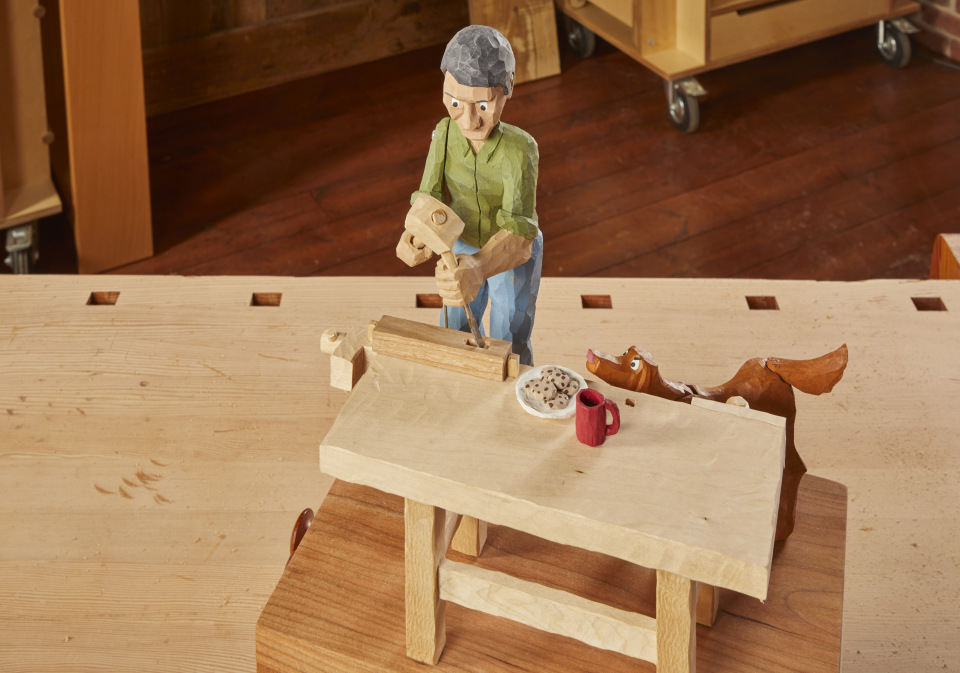 hand-carved person figurine of man with grey hair, green shirt, and blue jeans using mallet and chisel on wood. Next to him is a brown dog figurine, and figurines of a red mug and chocolate chip cookies