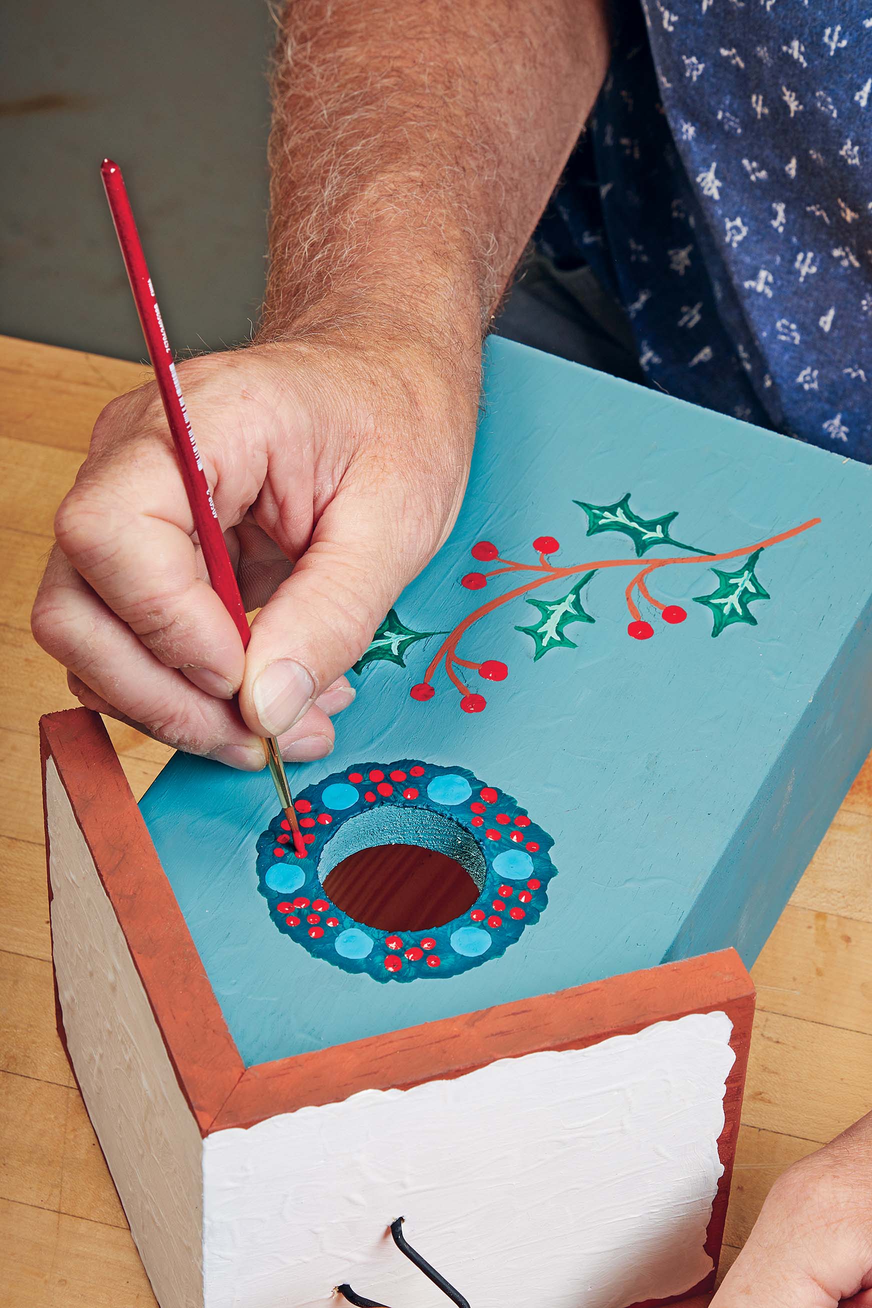 Using round brush to paint red dots for berries on a wreath aroud a bird house's hole