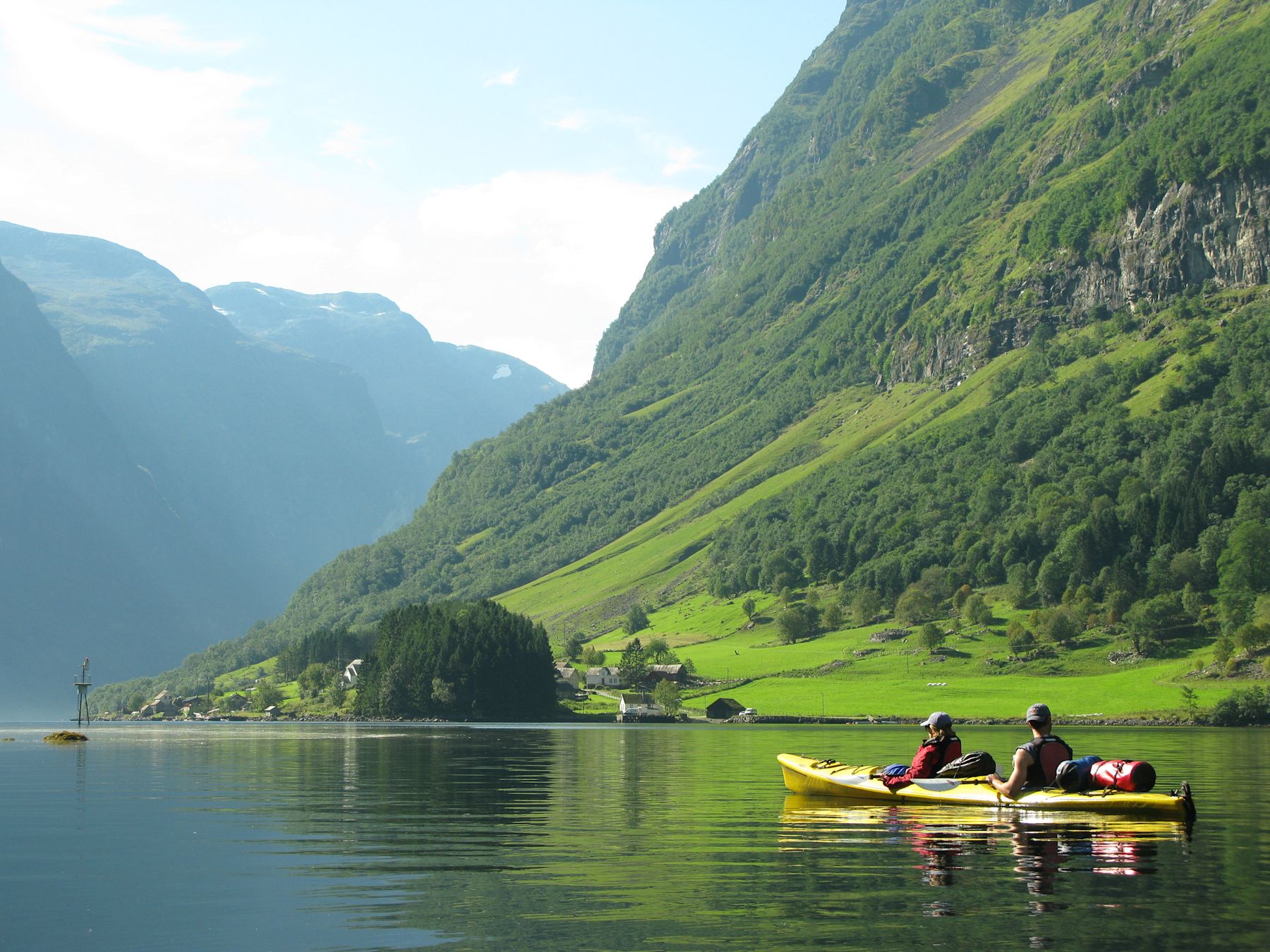 Two people paddling a yellow kayak across a still Norwegian fjord, below steep green mountainside