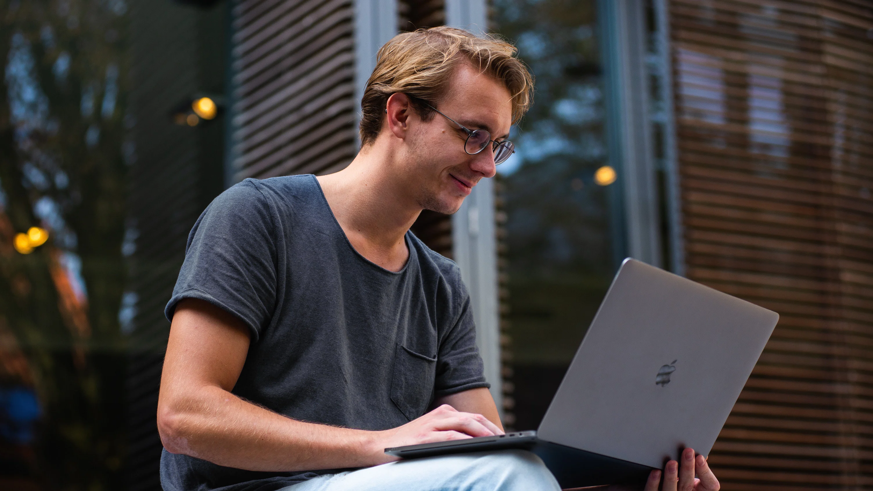 Young smiling man works on a laptop placed on his knees.