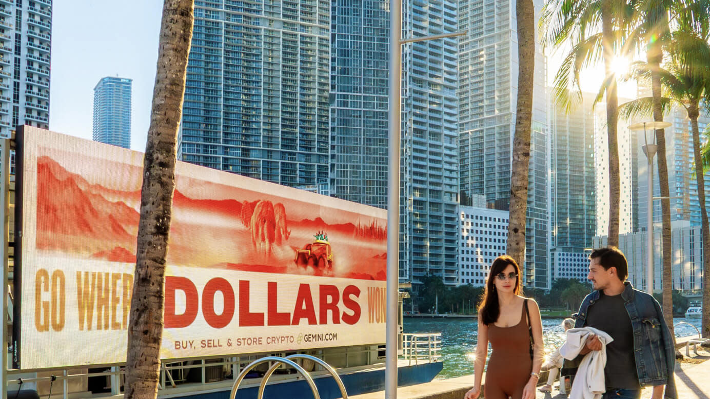 A couple walk by a Go Where Dollars Won't in a tropical city filled with skyscrapers