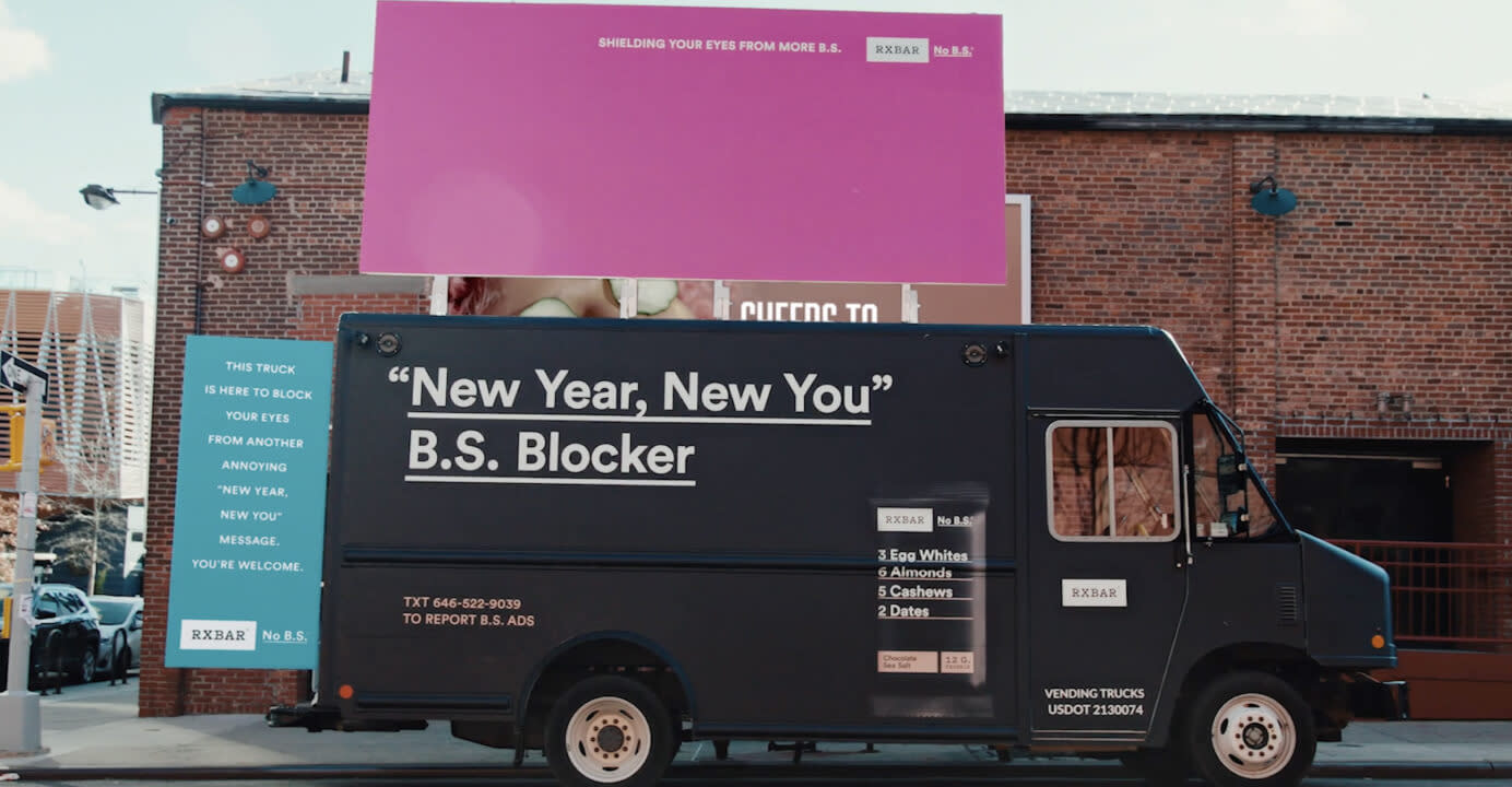 A black van with the writing 'New Year, New You B.S. Blocker' on it parked underneath a pink billboard with writing say 'shielding your eyes from more B.S.'