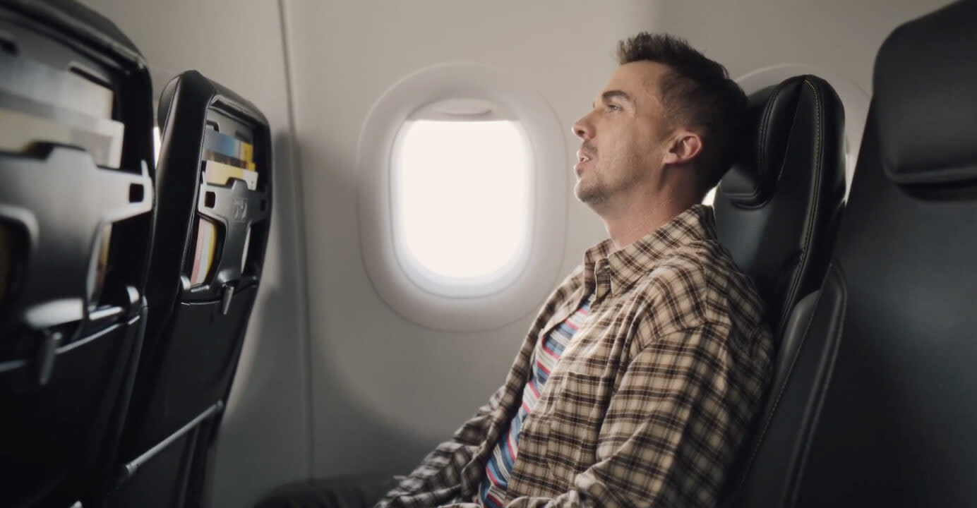 Frankie Muniz dressed in a plaid shirt sitting in a relaxed position on an airplane row's window seat