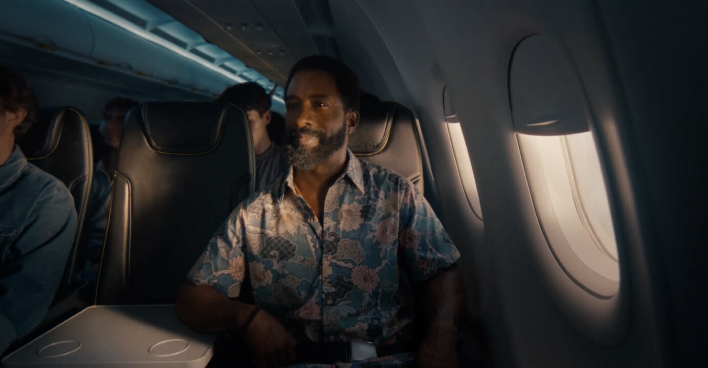 Man in a flowery shirt sitting in a Spirit airplane with the window shades half open with sunlight peaking in