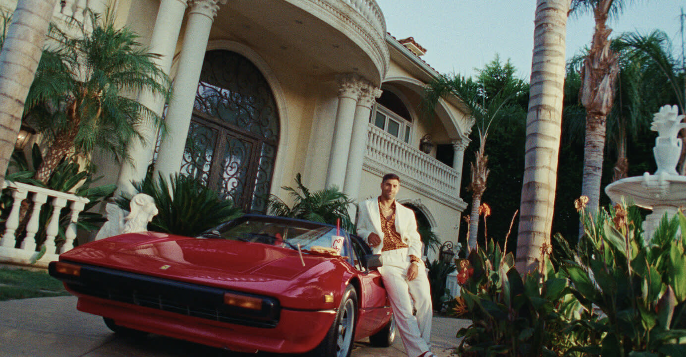 Jimmy G, a loaded Italian, standing next to a classic red sports car in front of an Italian villa inspired mansion