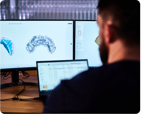 A person sitting at a desk with a laptop and two monitors displaying 3D models and files of dental scans.