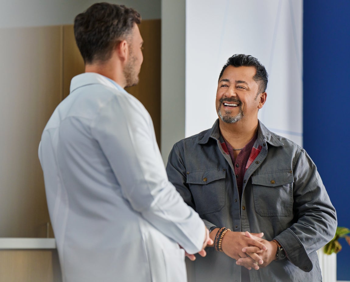 A male doctor in a white coat talks to a smiling male patient wearing a gray jacket in a clinical setting.