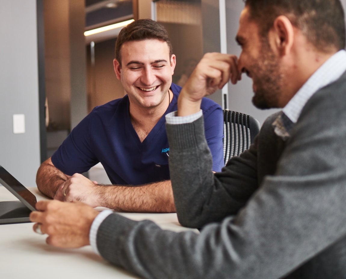 Two men sit at a table, one in dark blue scrubs and the other in a gray sweater. They are engaged in a conversation and smiling while looking at a tablet.