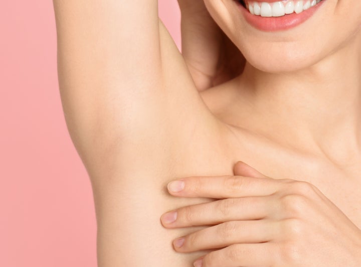 A woman is smiling and touching her armpit against a pink background