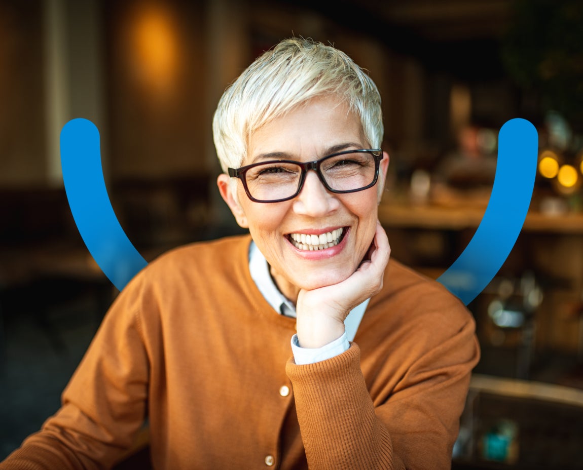 A smiling person with short, light hair and glasses, wearing an orange sweater, sits with one hand under their chin in a warmly lit indoor setting. Blue U-shaped design element is overlaid depicting a smile.