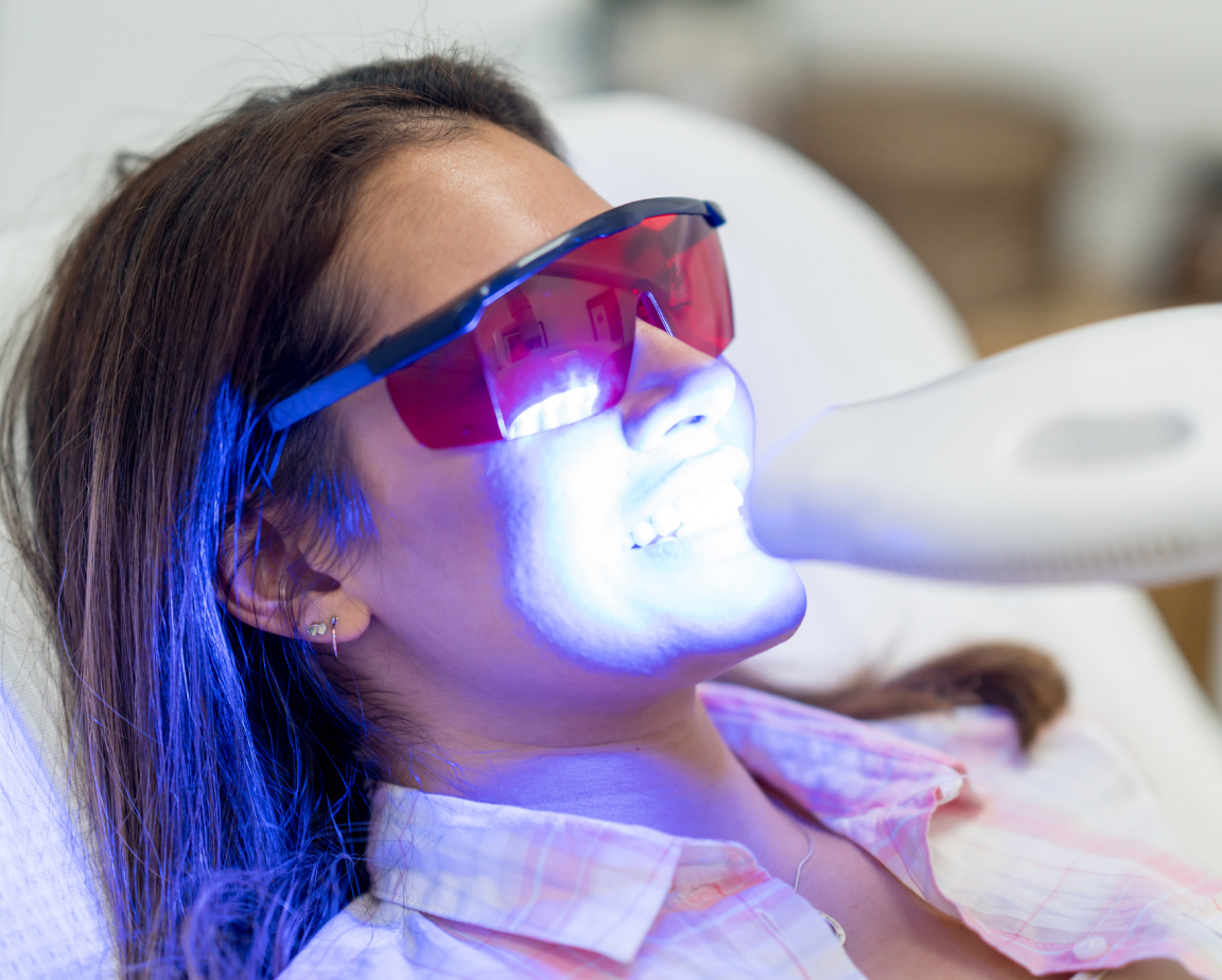 A woman wearing red protective glasses receives a teeth whitening treatment with a blue light at a dental office.