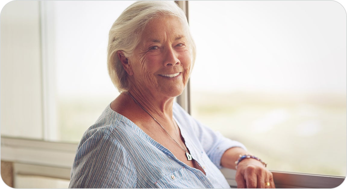 A female patient in a blue top, standing next to a window and smiling with her new dental implants after her gums have healed and fully recovered.