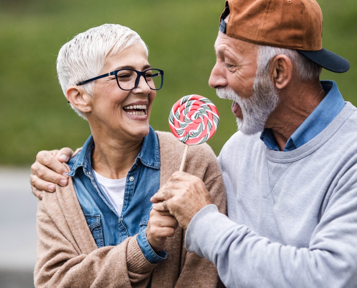 Smiling senior couple sharing a playful moment with a large lollipop, symbolizing joy and good dental health. Aspen Dental supports lasting smiles and oral wellness for all ages.