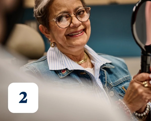 An older woman with glasses and a denim jacket smiles while holding a handheld mirror. A number '2' icon is visible in the bottom left corner.