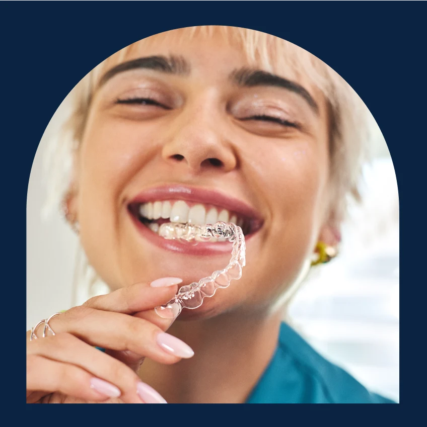 A person with light-colored hair smiles while holding a clear aligners near their mouth.